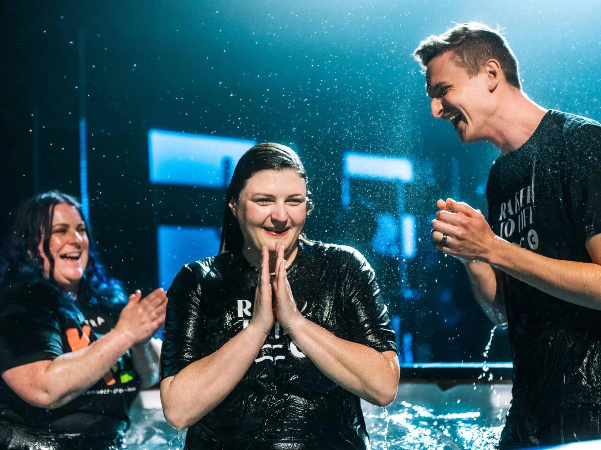A woman smiles with her hands pressed together in the baptism tank, drenched, as two people celebrate beside her.
