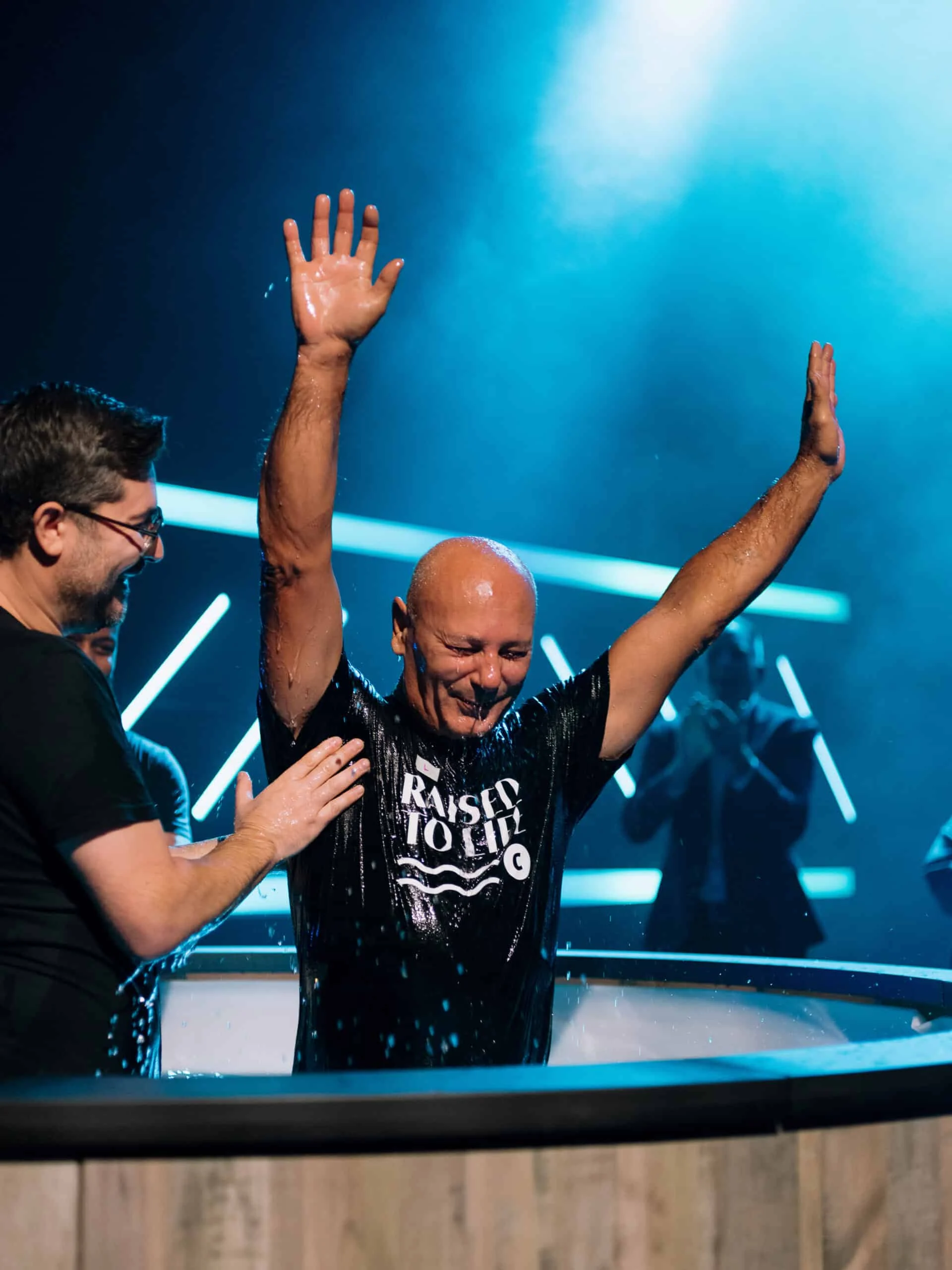 A man raises both arms in the baptism tank, water streaming off him, as a pastor rests a hand on his shoulder.