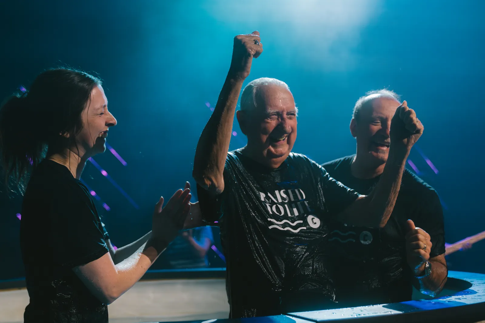 A man raising his fist and laughing moments after being baptized, flanked by two people celebrating with him at the baptism tank.