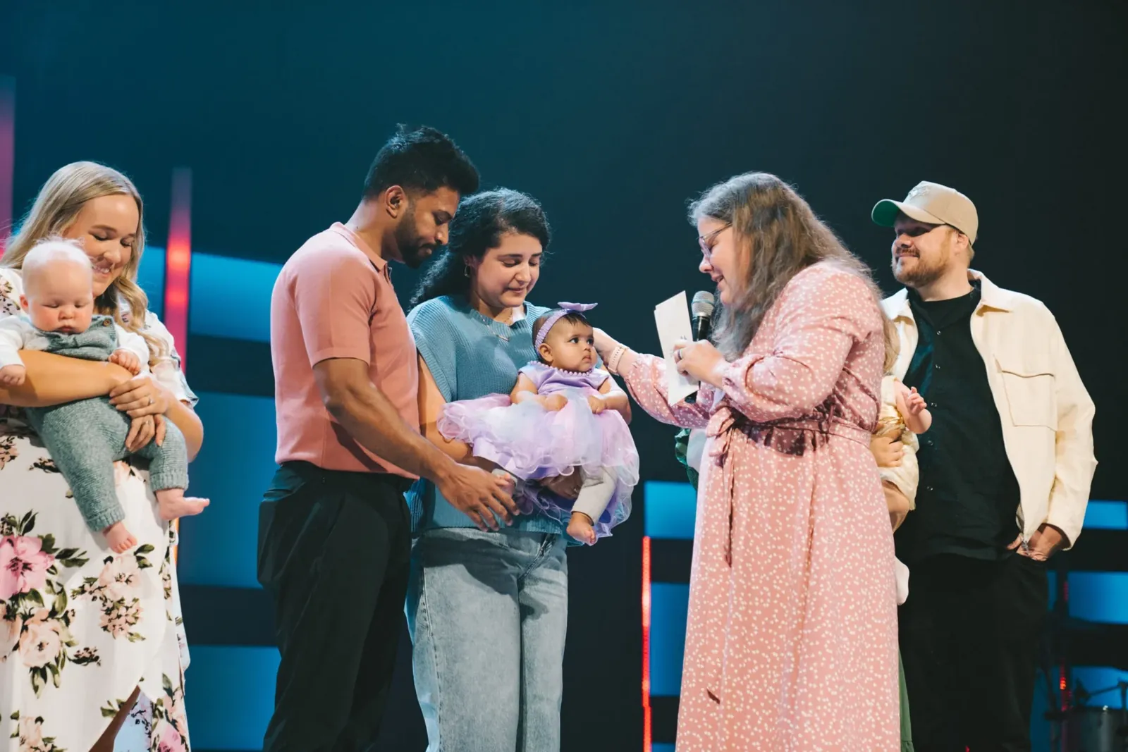 A pastor praying over a baby held by her parents on stage during a child dedication, with other families holding their children alongside.