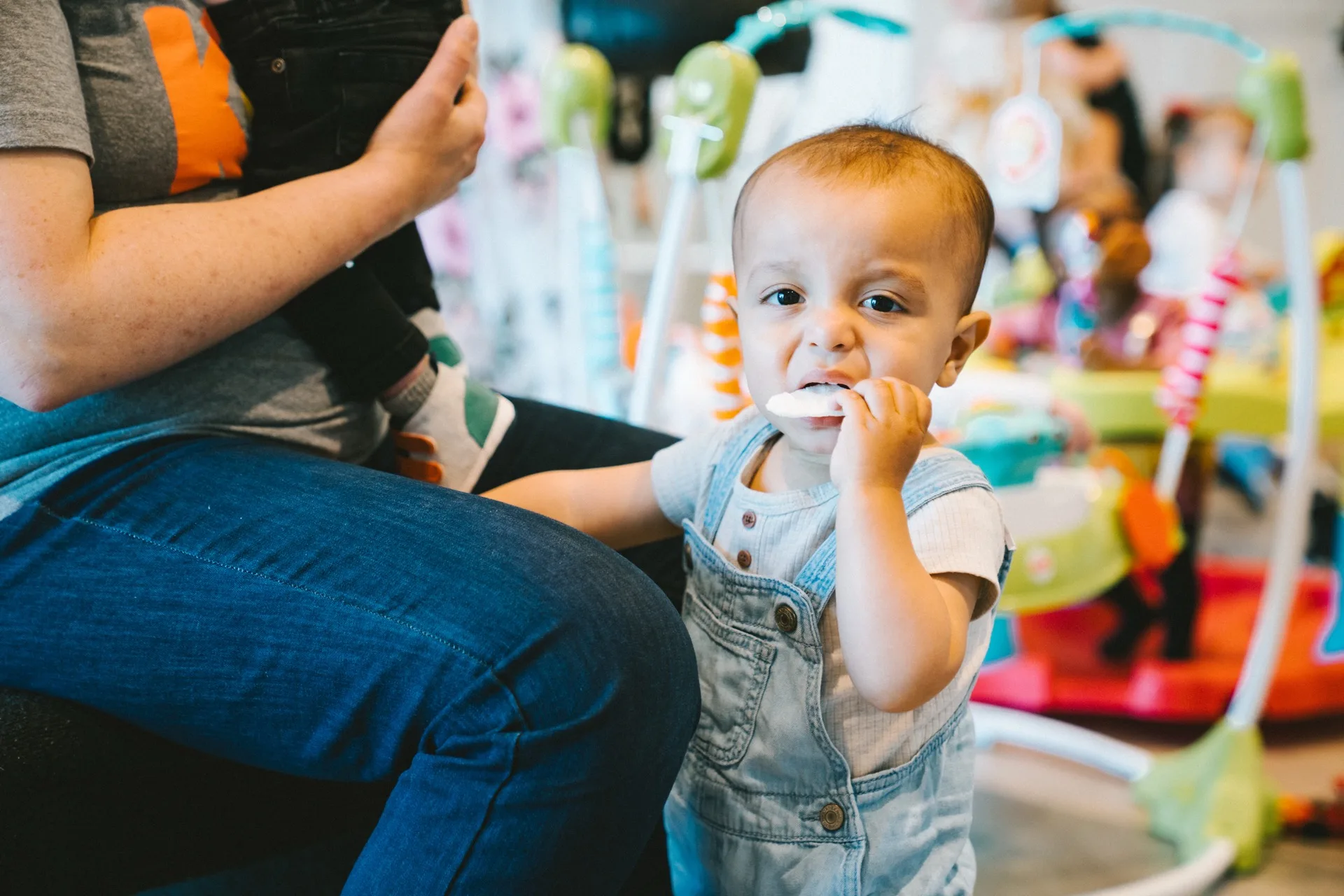 A baby standing beside an adult in a childcare room with toys and activity centres.