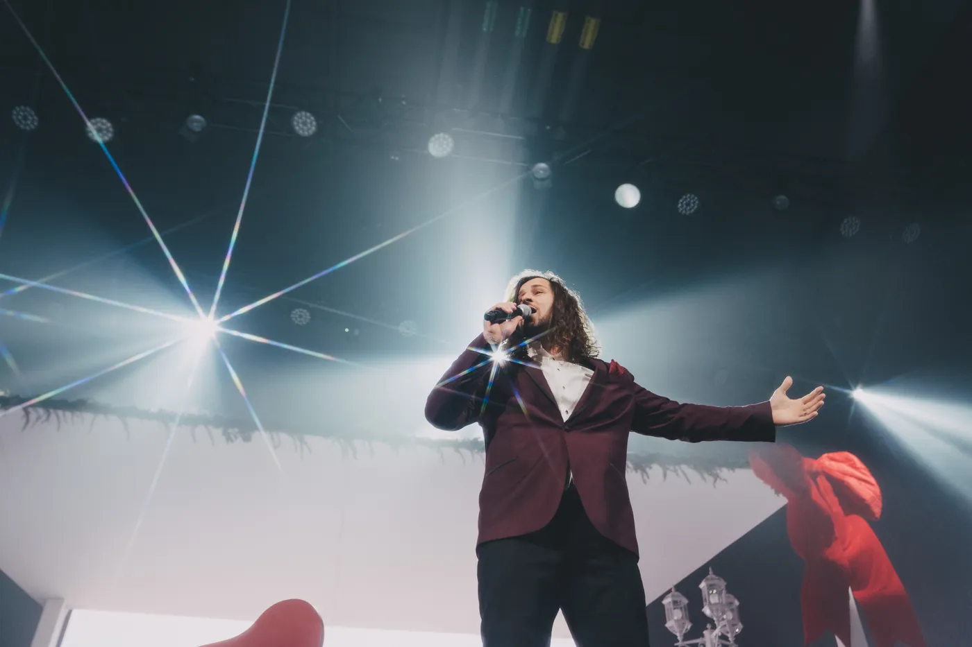 A singer in a burgundy jacket, arms open, backlit by starburst stage lights.
