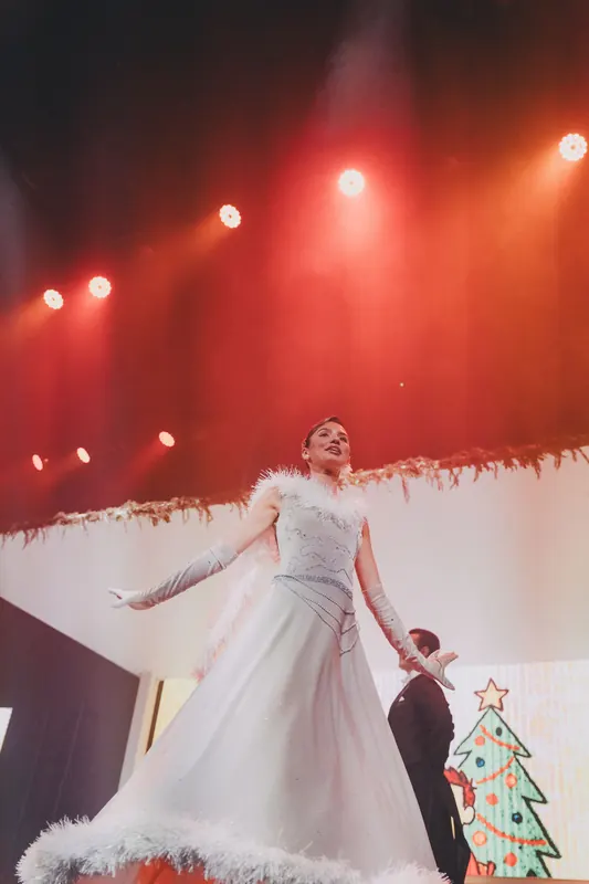 A dancer in a white gown moves across the stage beside a Christmas tree.