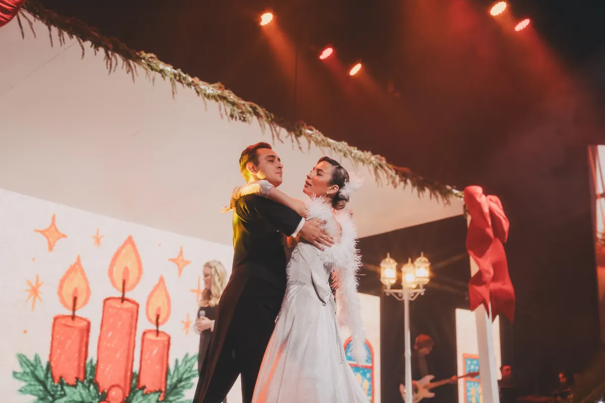 A couple in formal dress dance together in front of a painted backdrop of candles and holly.