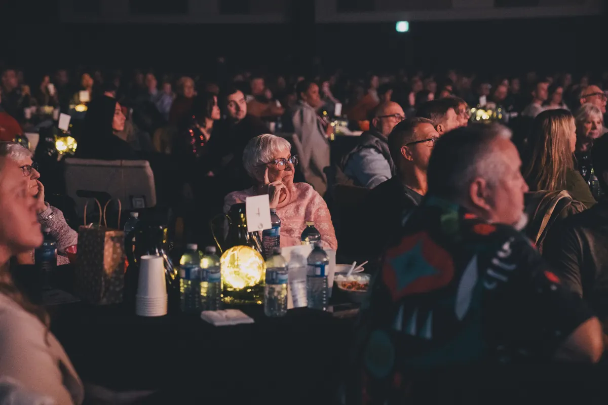 A full room of guests at candlelit tables, all turned toward the stage.
