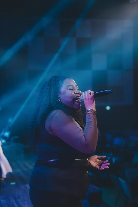 A vocalist sings into a handheld microphone, eyes closed, in blue light.