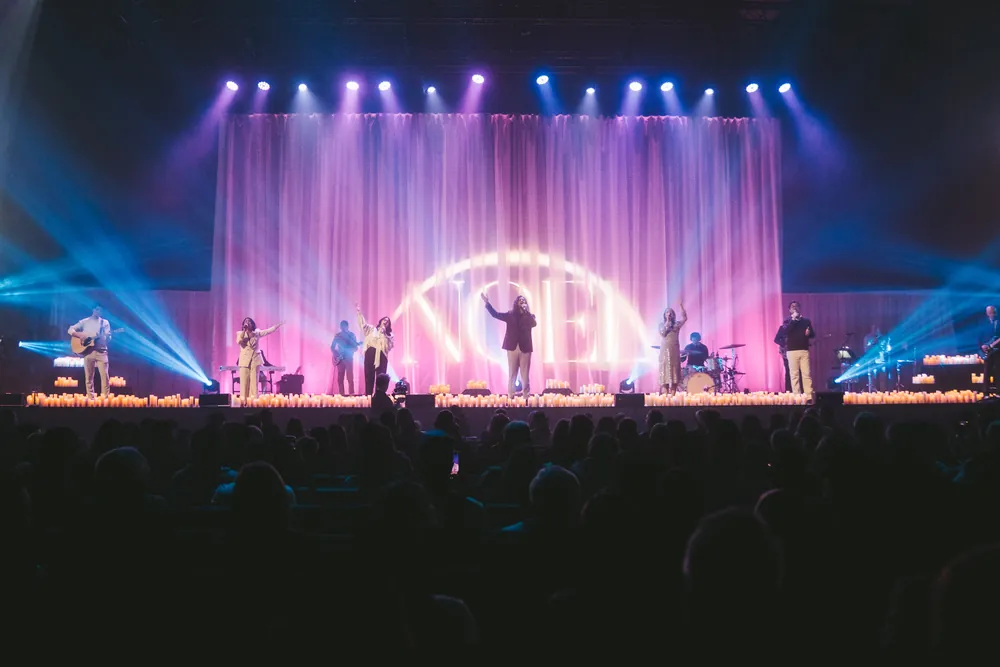 The band plays beneath a glowing NOEL sign on Christmas Eve, candles along the front of the stage.