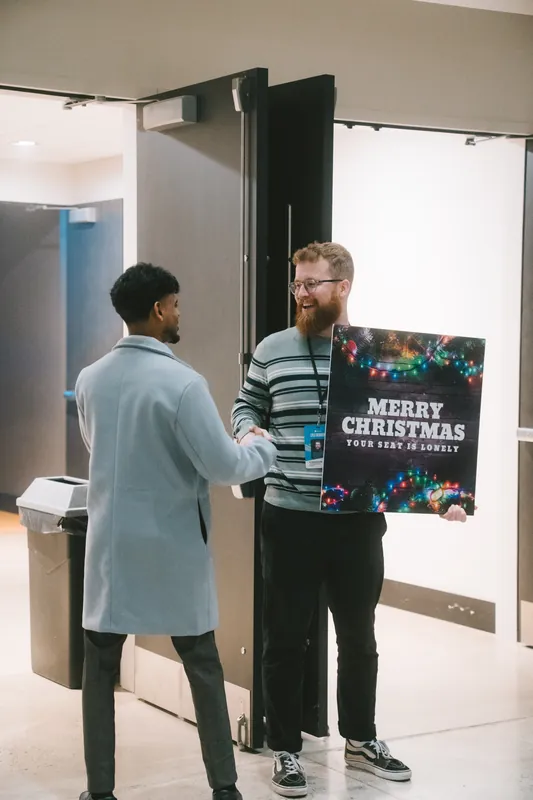 A greeter holding a Merry Christmas sign trimmed with string lights shakes hands with a guest at the doors on Christmas Eve.