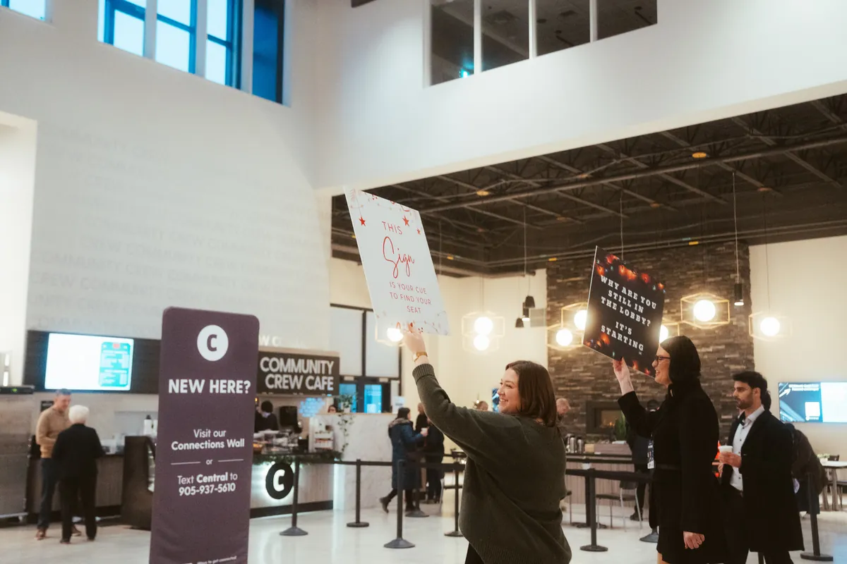 Smiling greeters in the lobby hold up playful signs on Christmas Eve, one reading &ldquo;This sign is your cue to find your seat.&rdquo;