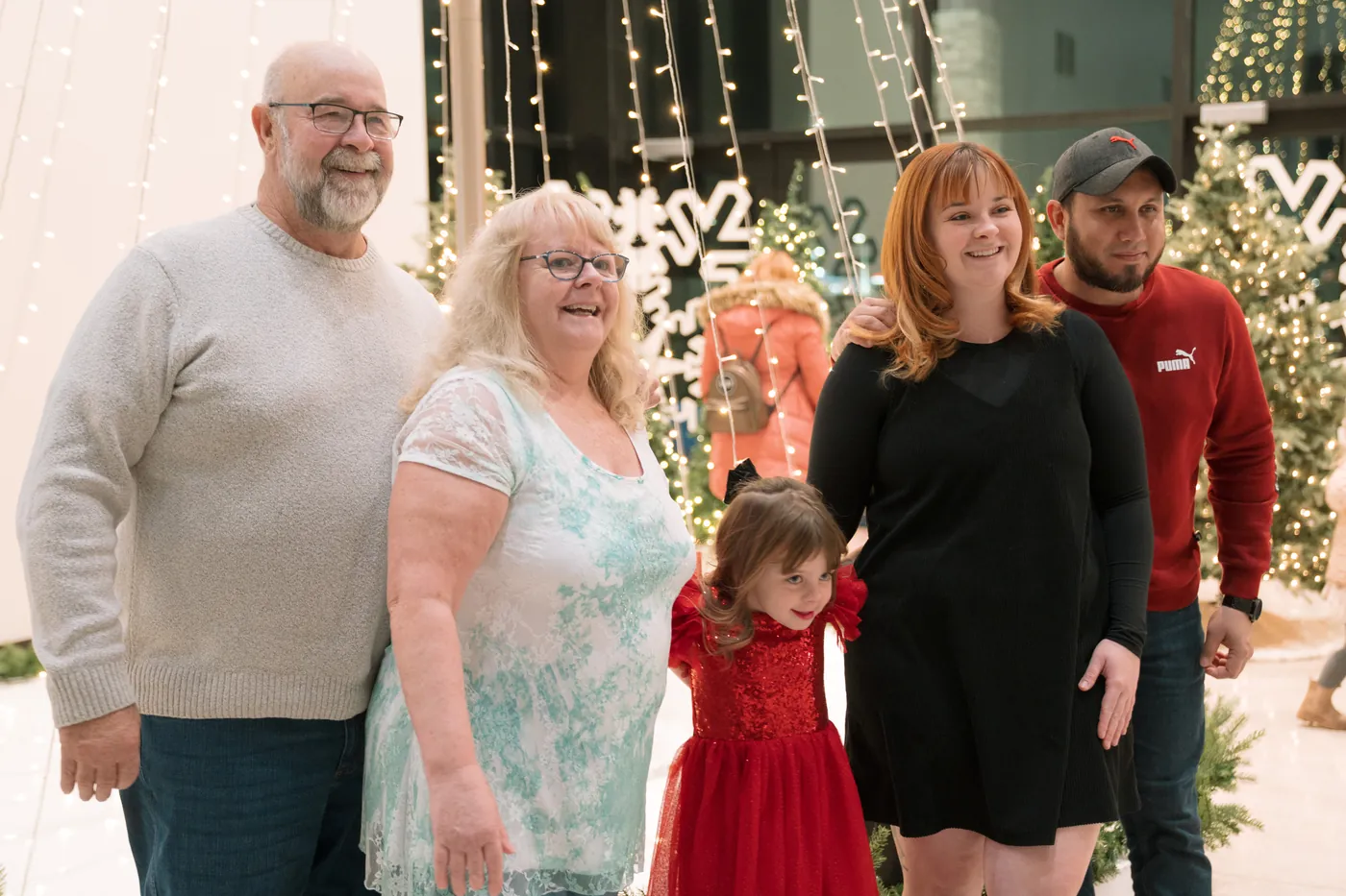 An older couple, a young family and a little girl in a red dress smile together among the lobby's Christmas lights.