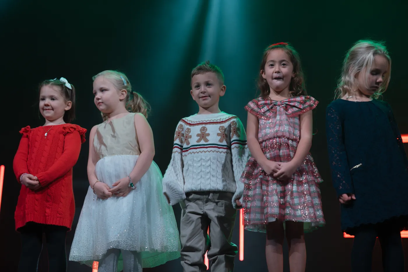 Five young children in Christmas dresses and sweaters stand together on stage under teal light.