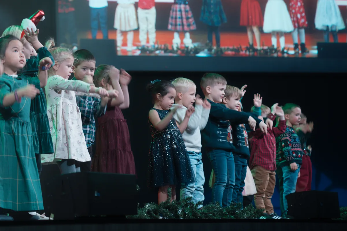 A row of children in Christmas outfits sing on stage, the big screen behind them showing the same moment.