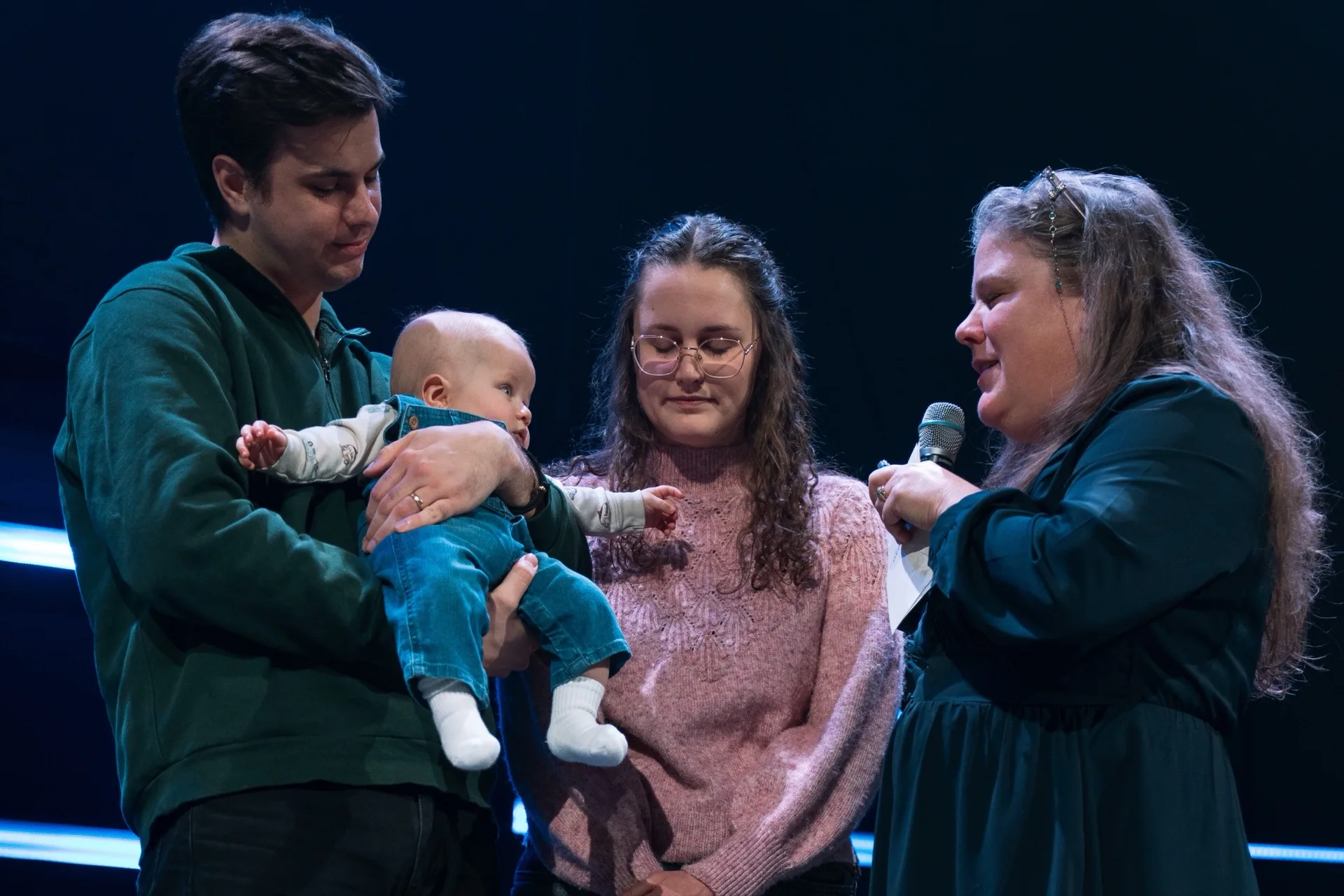 A couple holding their baby on stage during a child dedication, with a pastor beside them.