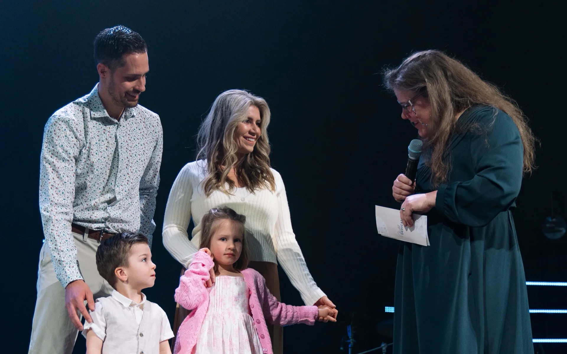 A family with two young children standing on stage with a pastor during a child dedication.