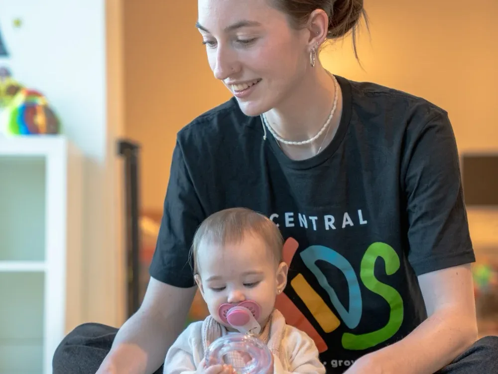 A Central Kids volunteer sitting on the floor with a baby who is playing with a rattle.
