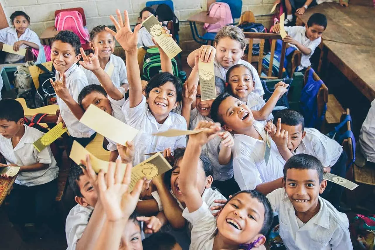A classroom full of smiling children waving and holding printed bookmarks toward the camera.