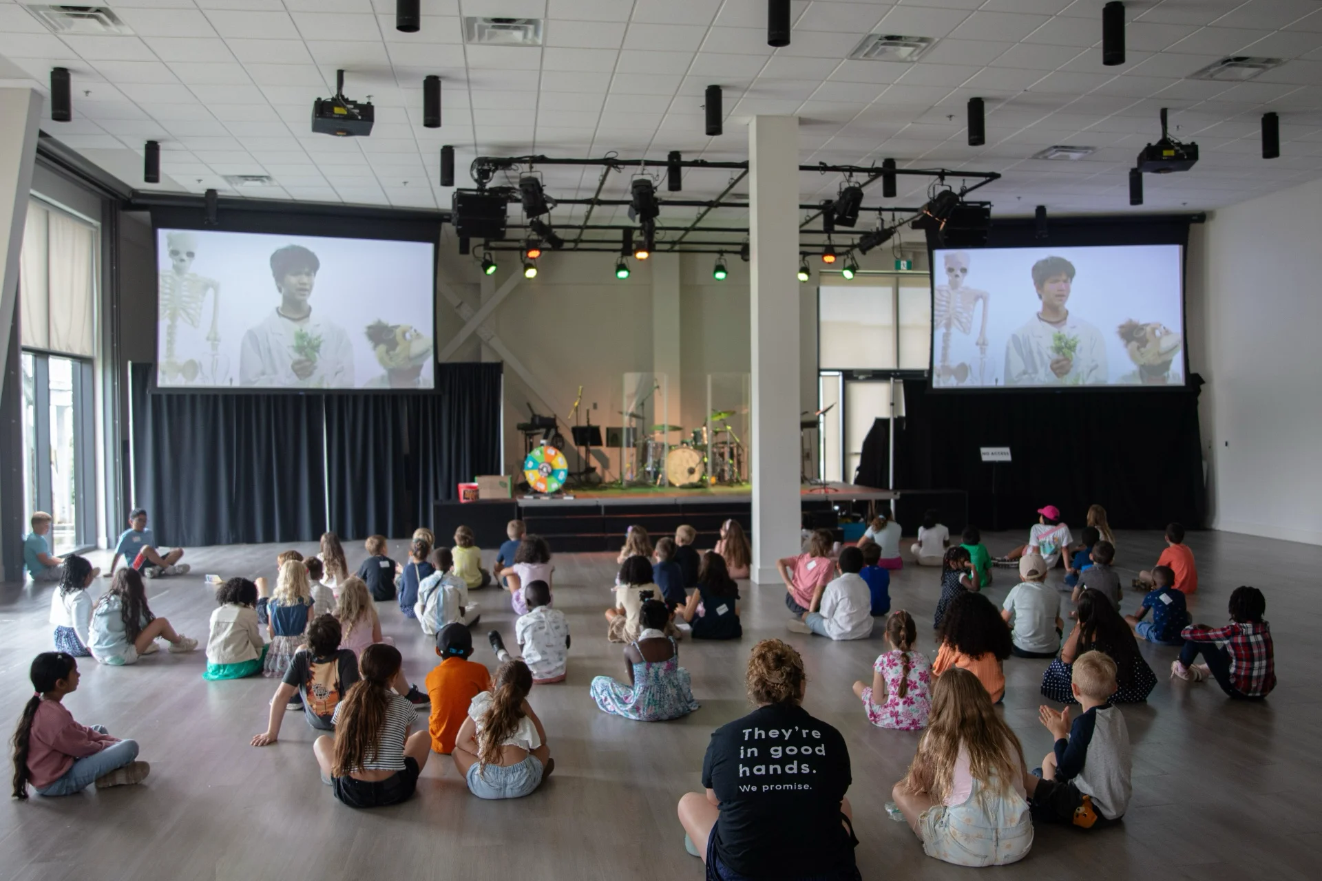 A large group of children sitting on the floor of a bright room, facing two screens and a stage during a kids programme.