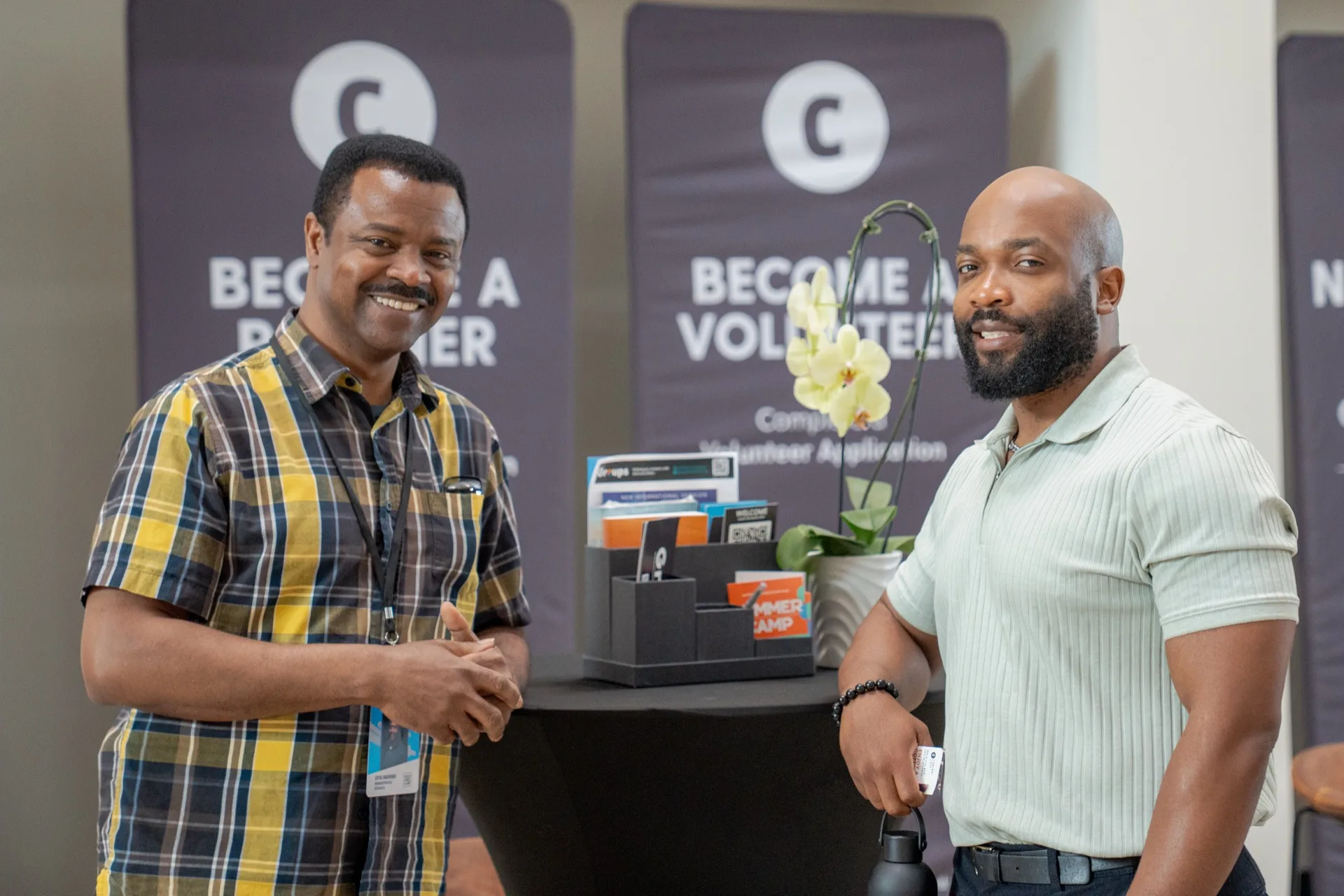 Two men standing and smiling in front of the Become a Partner and Become a Volunteer banners in the lobby.