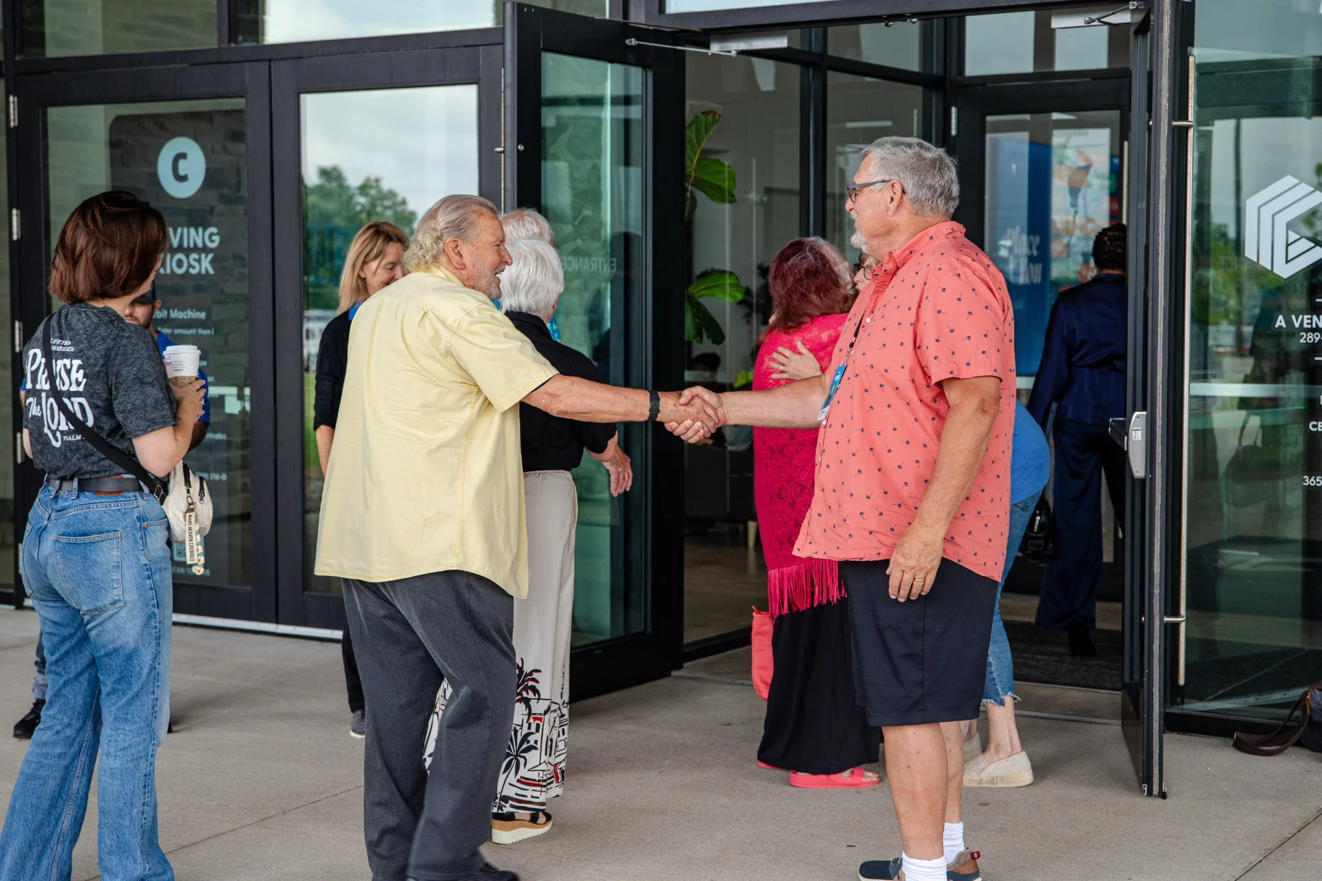 Two people shaking hands in the open glass front entrance of the building while others walk in, one carrying a coffee cup.