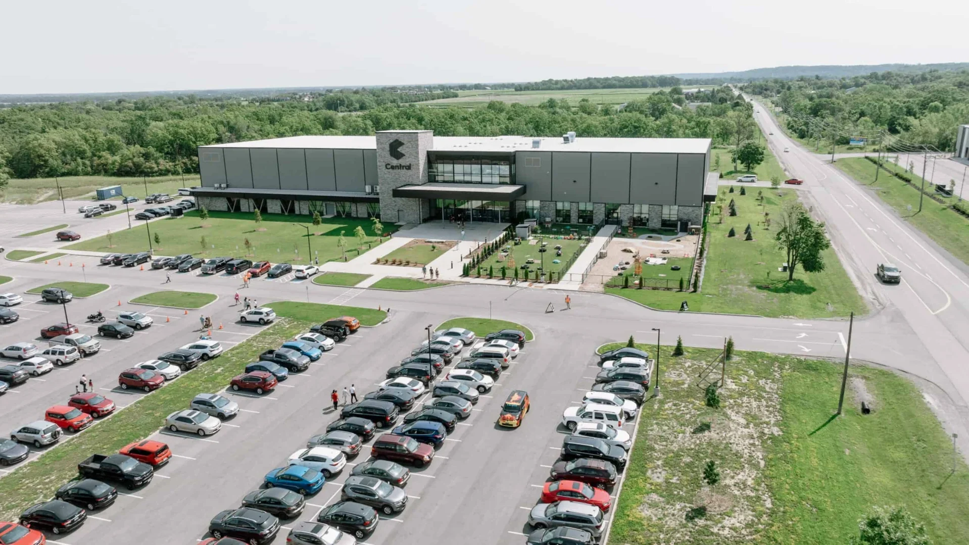 People walking from a full parking lot toward the Central building while a person in an orange safety vest stands near the drive aisle.