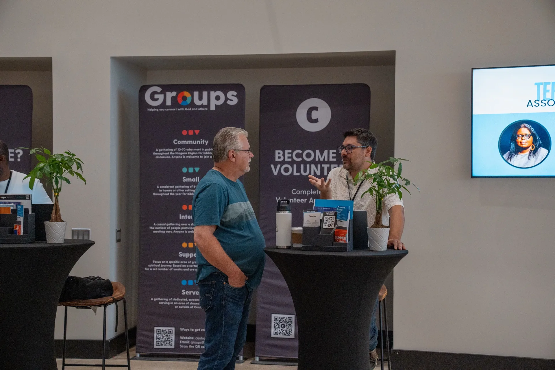 Two men talking beside the Groups and Become a Volunteer banners at the Connections wall.